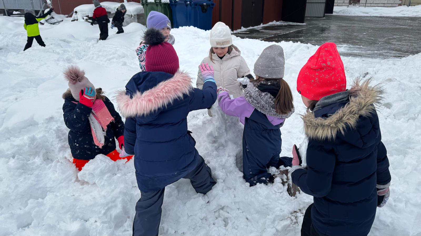 Grupa dzieci ubranych w zimowe ubrania bawi się i lepi bałwana na zaśnieżonym podwórku.
