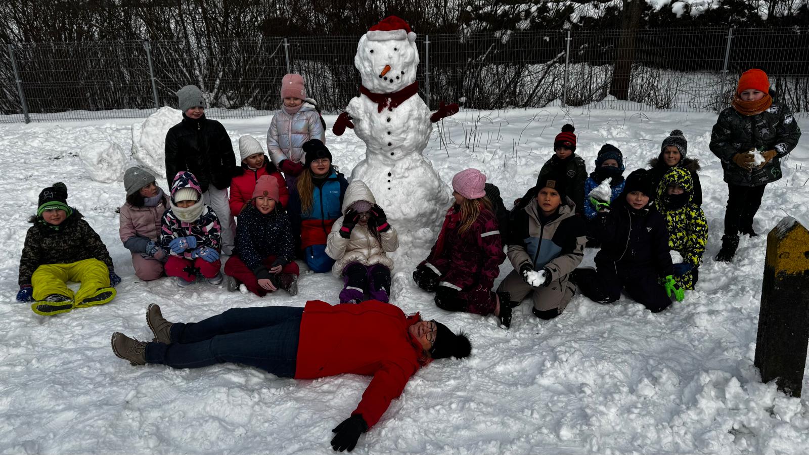 Grupa dzieci ubranych w zimowe ubrania bawi się i lepi bałwana na zaśnieżonym podwórku.